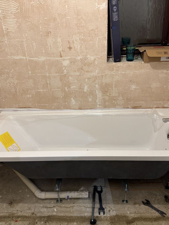 A bathroom stripped back to bare wall with a new bath in place on its cradle, pipework and tools on the floor