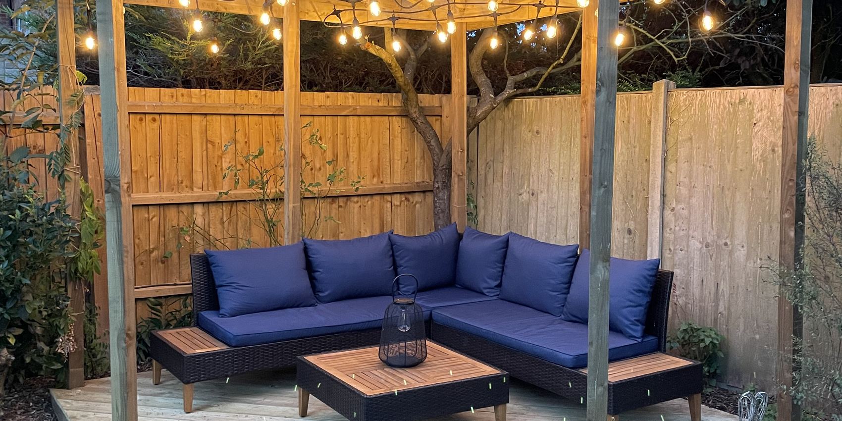 A timber pergola and deck lit with festoon lights at dusk, with a corner sofa beneath it in a Cambridgeshire garden