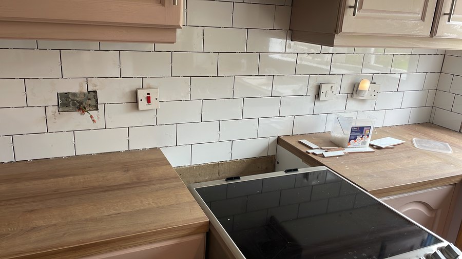 A kitchen part way through a refresh: white metro tiles being laid with spacers still in, new oak-effect worktops and hob fitted, one socket replaced and the next opened up