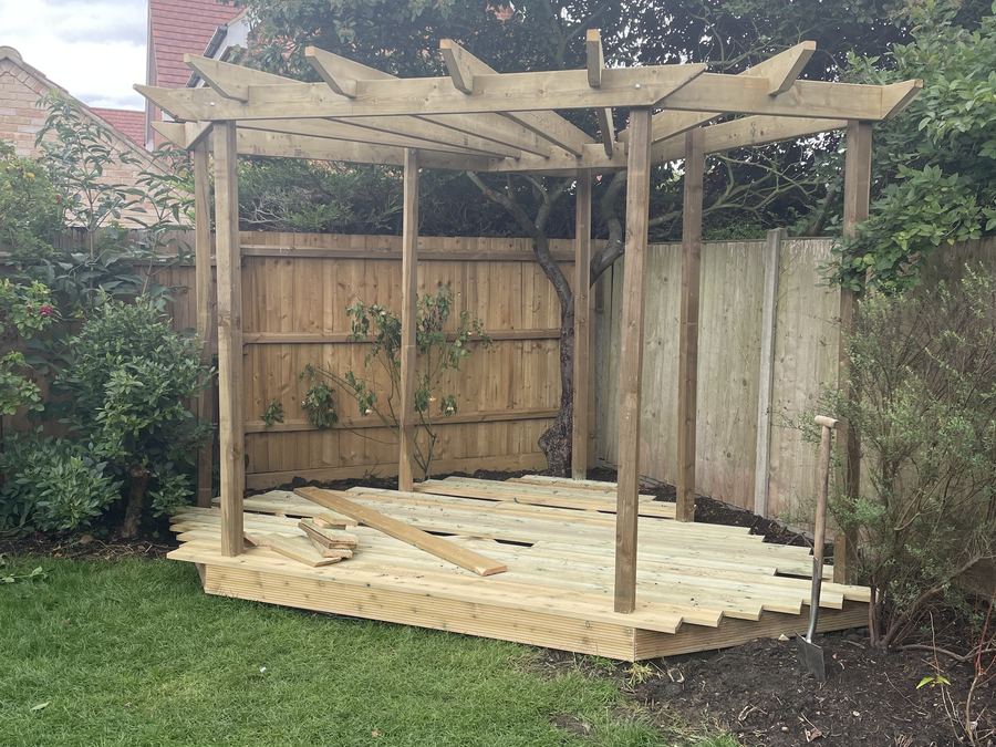 A completed timber pergola and deck in a garden, seen in daylight against a fence and planting