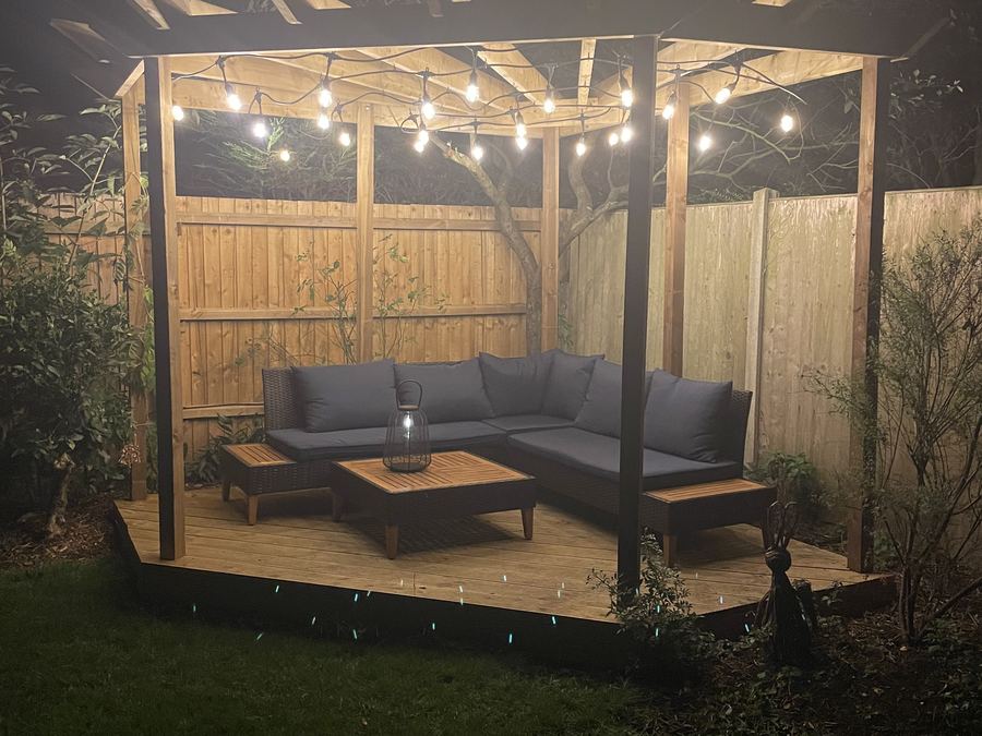 The finished pergola at dusk, lit with festoon lights above a corner sofa on the new deck